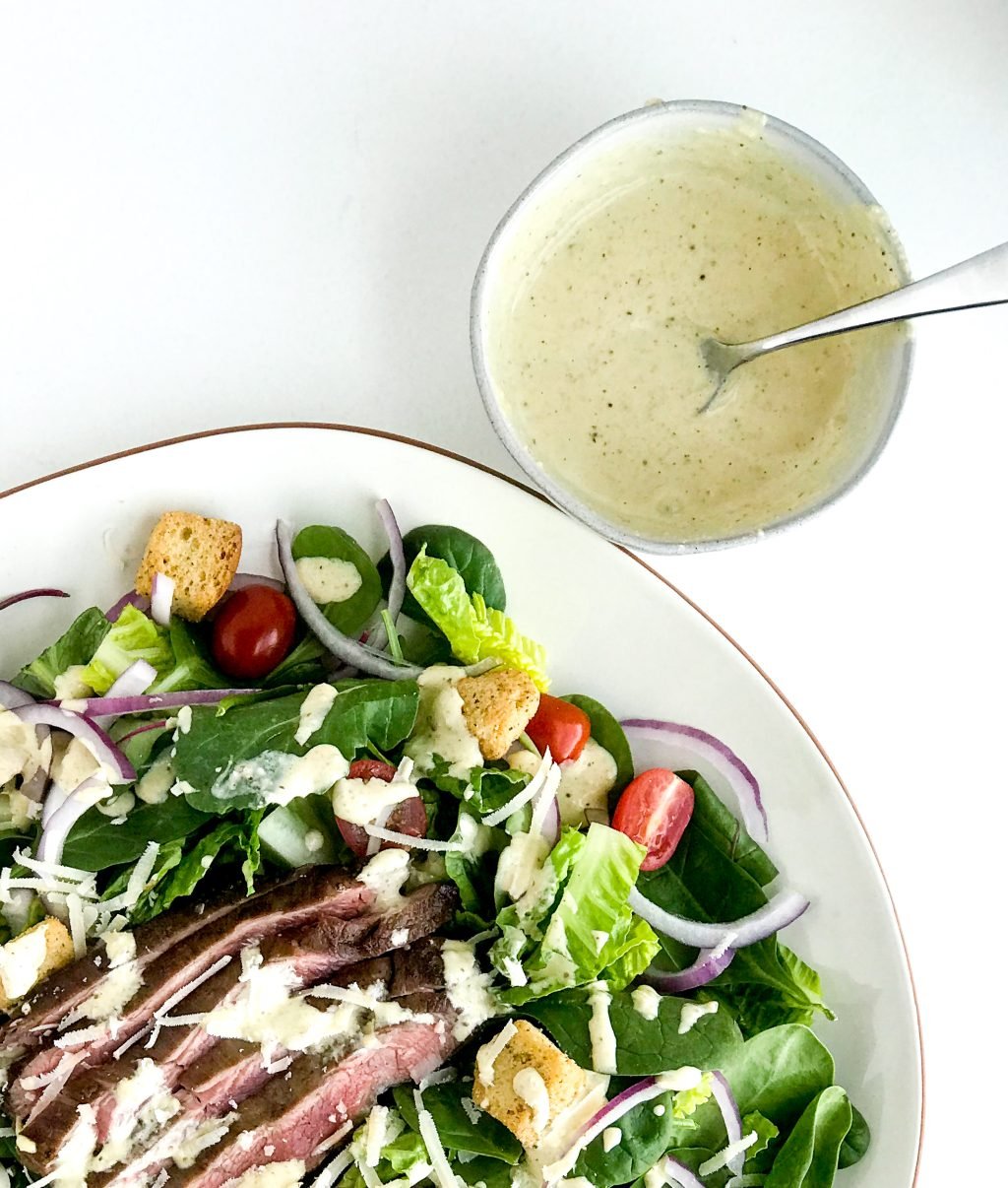 salad on a plate with caesar salad drizzled on top sitting next to a bowl of caesar dressing with a spoon inside.
