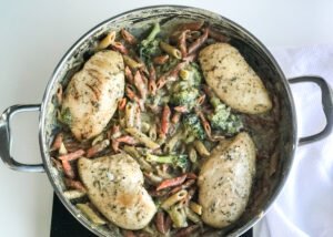 chicken breast in a pan with penne noodles, broccoli, and a cheesy roux